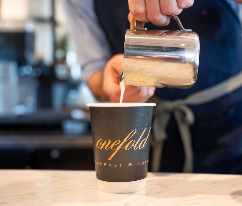 A barista pours steamed milk into an Onefold cup on a marble counter